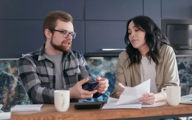 A couple sits at a kitchen table reviewing documents and discussing finances.