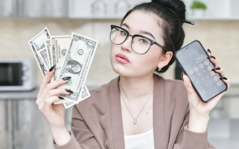 Woman holding cash and smartphone displaying calculator, highlighting personal finance management.