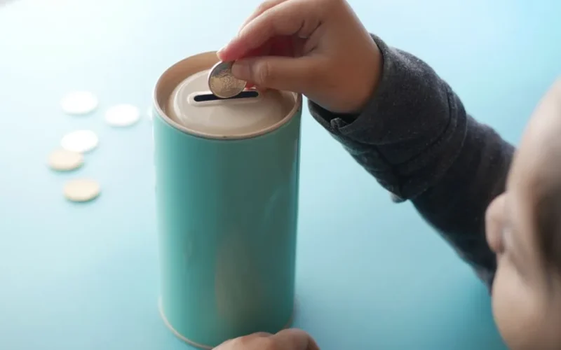 A child placing a coin into a teal piggy bank, representing saving money and financial education.