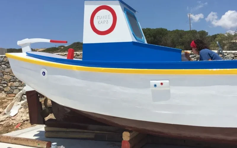 A vibrant fishing boat being painted by a woman in Naxos, Greece, under clear skies.