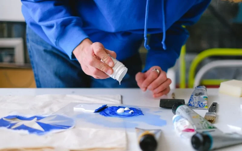 Unrecognizable male master with small jar adding white paint into blue pigment while working at table with stencil printing in studio on blurred background