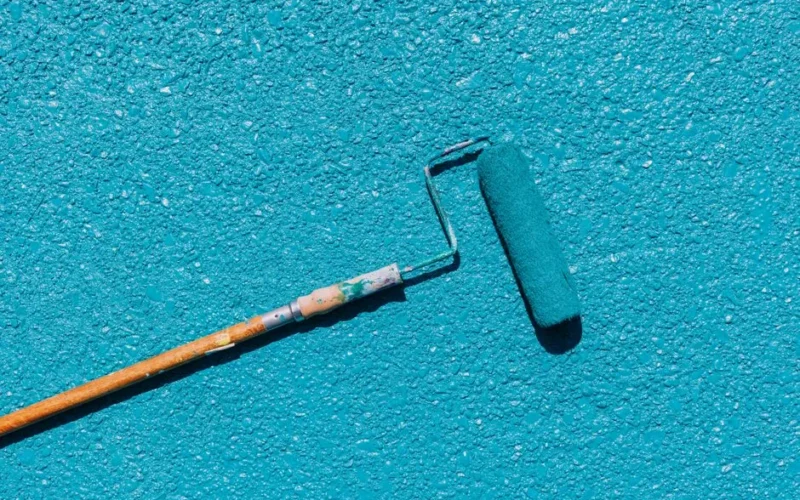A close-up of a blue paint roller applying paint on a textured wall surface, captured in bright sunlight.