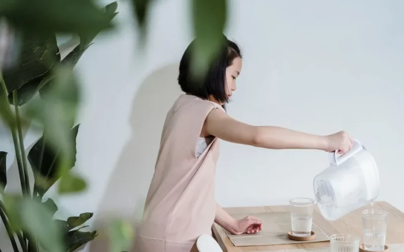 A young girl pours water from a pitcher into glasses at a wooden table indoors.