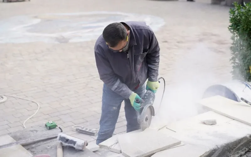 A construction worker uses a power saw to cut stone tiles outdoors, creating dust as he works.