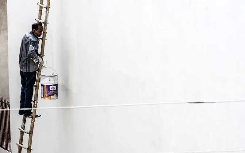 A worker on a ladder paints a large white wall outdoors, showcasing construction work.
