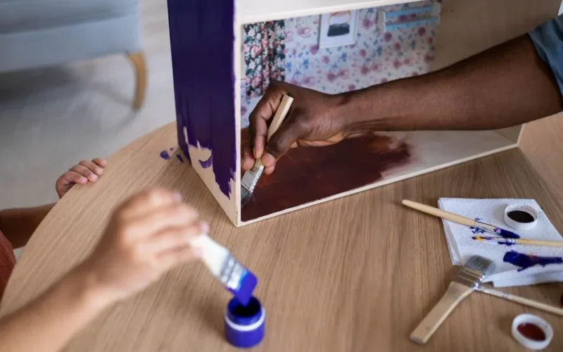 Adults and child creatively painting a wooden dollhouse using vibrant colors and brushes.