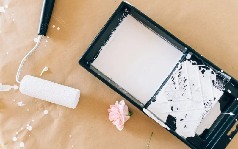 Close-up of a paint roller, tray with white paint, and a pink flower on craft paper.