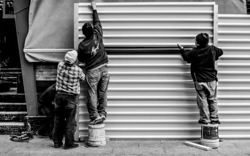 Black and white photo of three workers assembling metal panels outdoors, standing on buckets.
