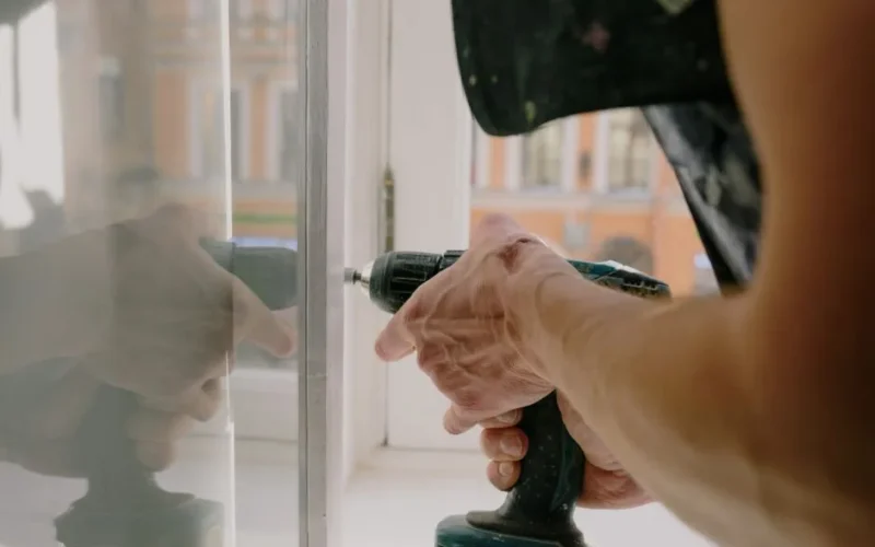 Unrecognizable professional male worker assembling white window with cordless screwdriver with drill near windowsill with reflection on glass during repair works in apartment