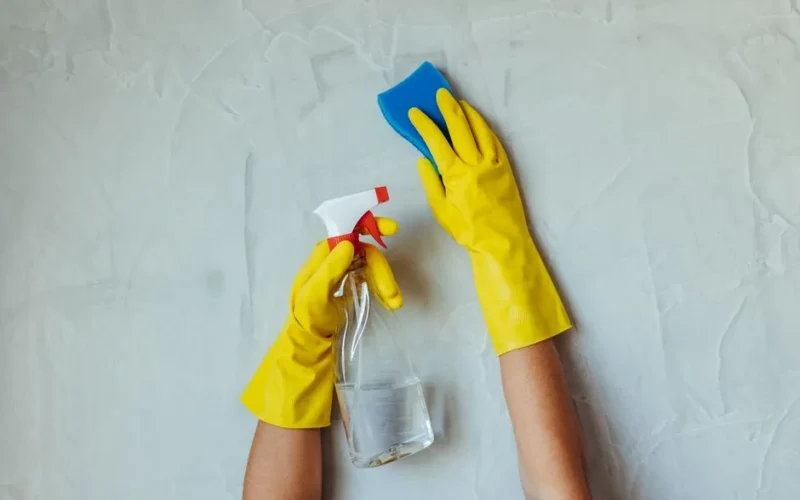 Close-up of hands in yellow gloves cleaning a wall with a spray bottle and sponge.