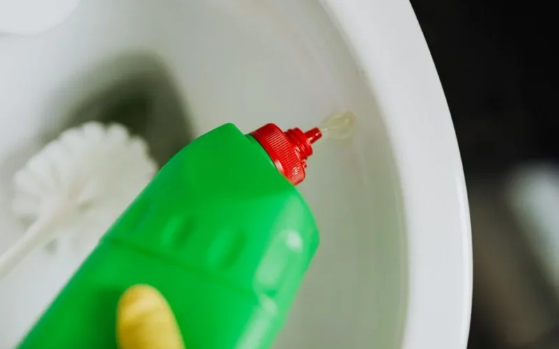 Top view of crop faceless person in yellow rubber gloves cleaning toilet with brush and pouring liquid disinfecting cleaner in toilet bowl