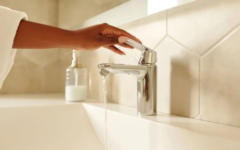 A close-up shot of a hand operating a chrome bathroom faucet with water flowing in a modern bathroom setting.