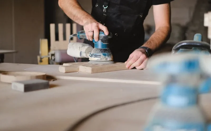 Unrecognizable male woodworker wearing uniform and polishing wooden board with random orbital sander at big table in professional studio