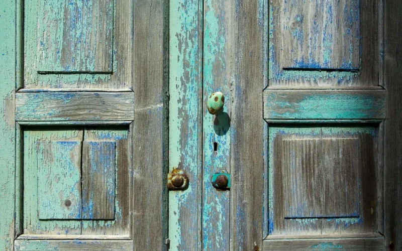 Close-up of weathered wooden doors showcasing rustic blue and turquoise paint texture with vintage doorknob.