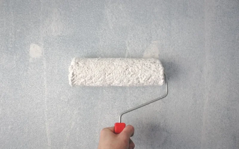 A person's hand using a paint roller to apply white paint on a wall.
