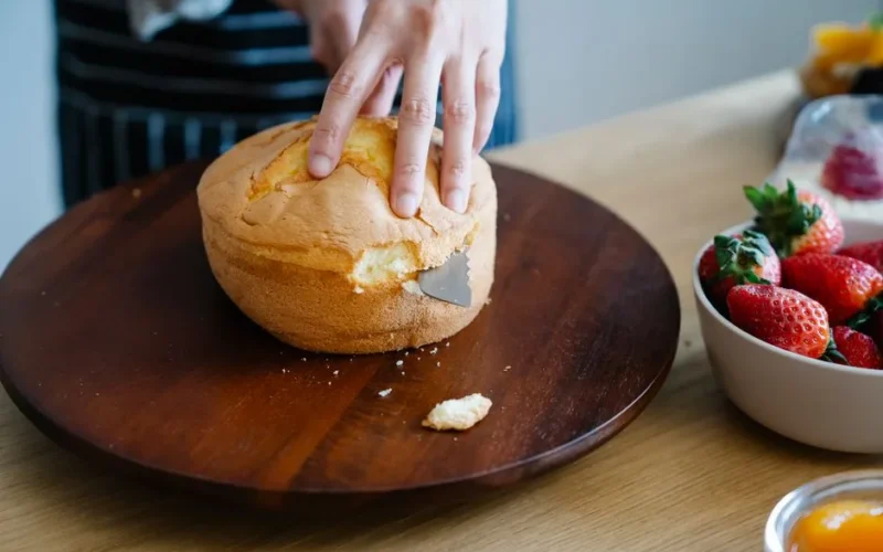 Hand slicing a sponge cake on a wooden platter with fresh strawberries nearby.