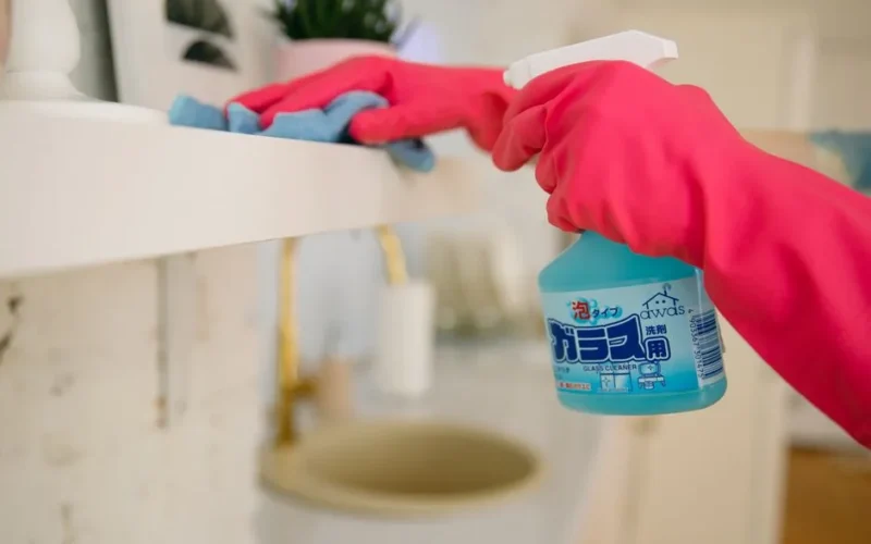 A woman using a blue spray cleaner and cloth while wearing pink gloves for home cleaning.