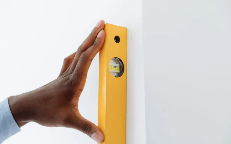A close-up of a hand using a yellow level tool against a white wall to ensure vertical alignment.
