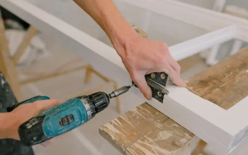 Unrecognizable professional male master using cordless screwdriver to install metal hinge on wooden window in light room during repair works