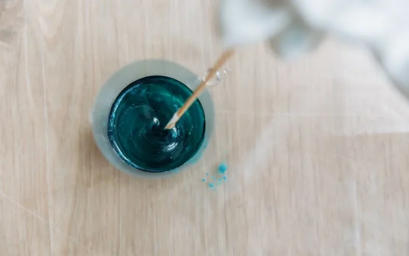 Top view of vibrant blue paint being mixed with a stick in a clear bowl on a wooden table.