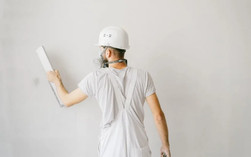 Back view of a construction worker in safety gear plastering a white wall.
