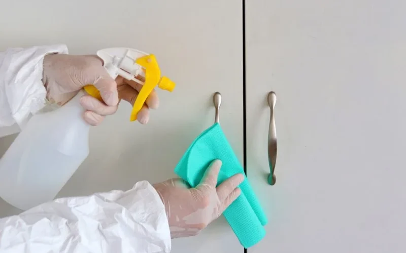 Gloved hands cleaning cabinet handles with a spray bottle and cloth for hygiene.