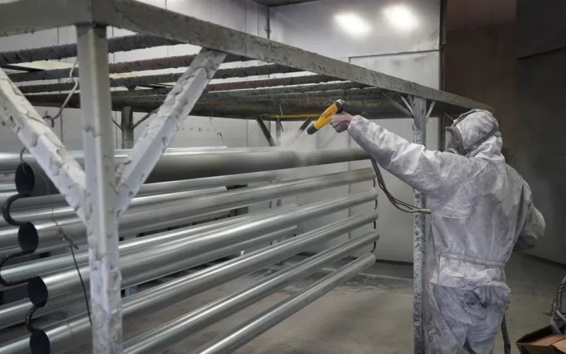 Worker in protective gear applying powder coating to metal pipes in a factory setting.