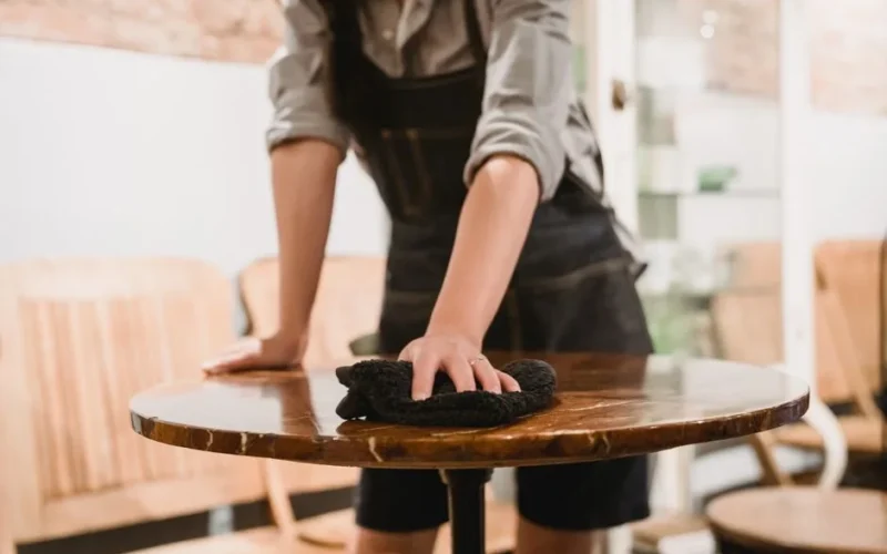 Person in apron cleaning a wooden table in a cozy café environment, focusing on hygiene.