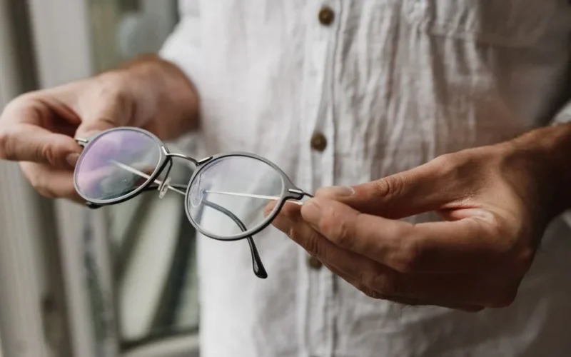 A man holds eyeglasses in a close-up shot, showcasing a casual shirt indoors.