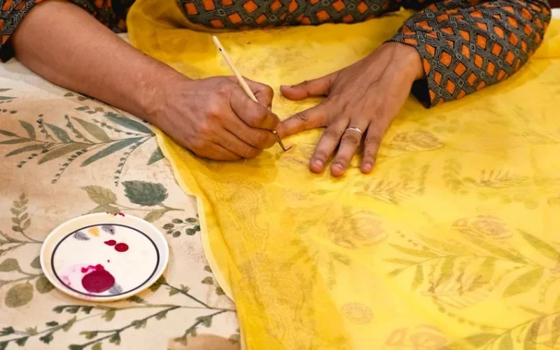 Artisan painting intricate design on yellow fabric with brush.