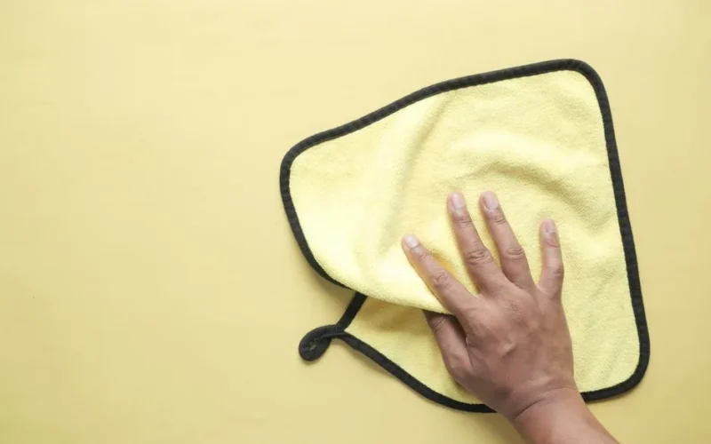 A hand using a microfiber cloth to wipe a yellow background, demonstrating cleaning and hygiene concepts.