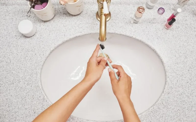 Close-up of hands cleaning a toothbrush under running water in a modern bathroom.