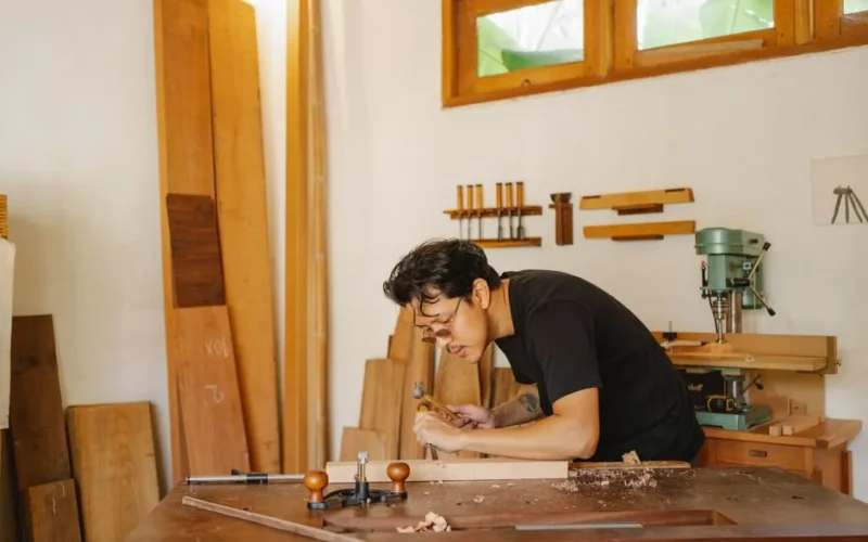 Side view of concentrated Asian craftsman creating wooden plank with hammer and chisel during work in professional joinery
