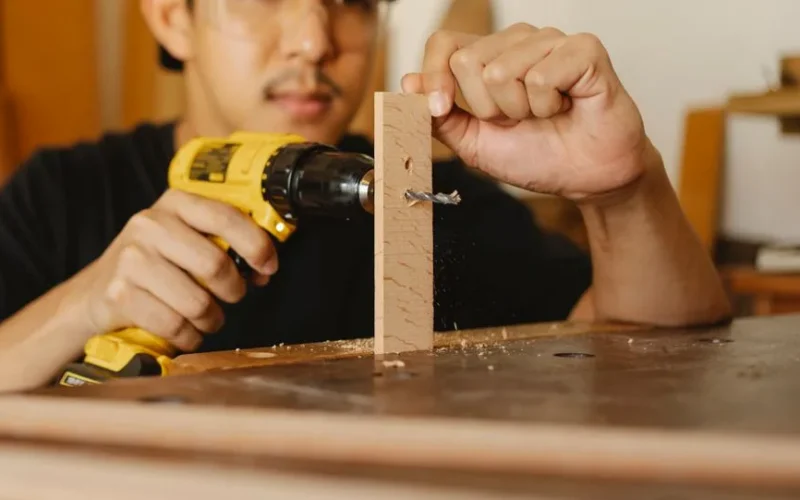 Crop anonymous ethnic male drilling timber detail for making holes while working on table in workshop