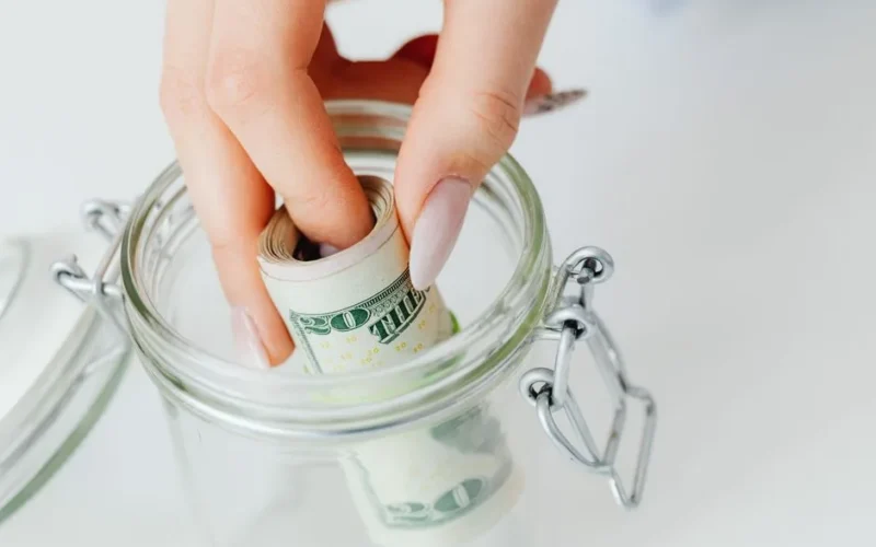 A close-up of a hand placing rolled dollars into a glass jar, symbolizing savings.