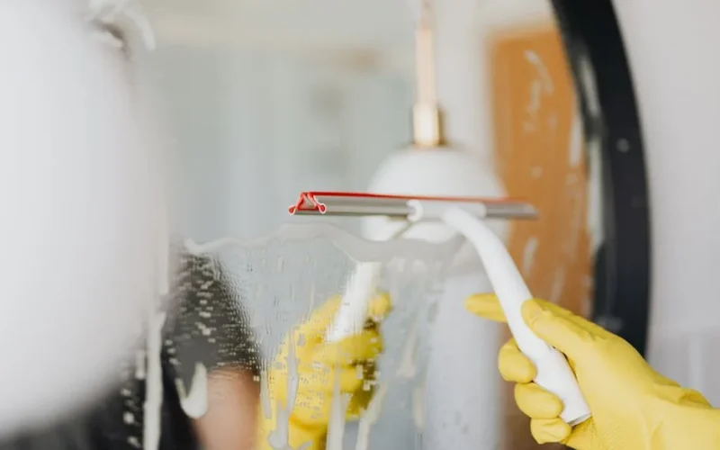 Anonymous person in yellow rubber gloves wiping water by squeegee from mirror in bathroom