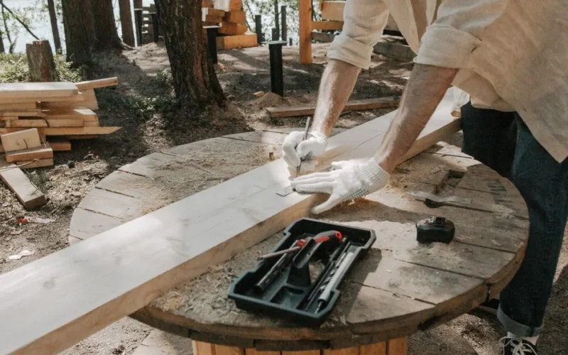 A carpenter skillfully outlines on a wooden plank with tools outdoors, surrounded by trees.