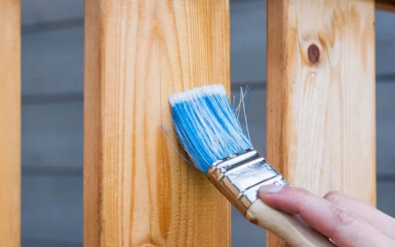 A hand painting a wooden fence plank with a blue paintbrush outdoors. Perfect for DIY and renovation themes.