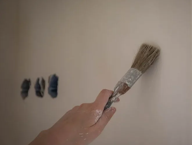 A close-up of a hand using a brush to paint a white wall, three color swatches visible.