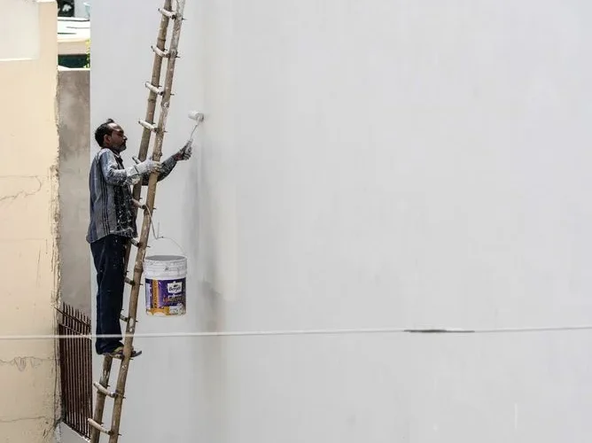 A man on a ladder painting a building's exterior wall with a roller brush outdoors.
