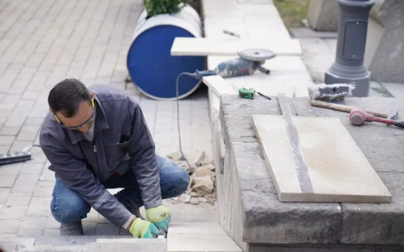 A focused mason wearing protective gear, laying stone tiles outdoors, showcasing craftsmanship.