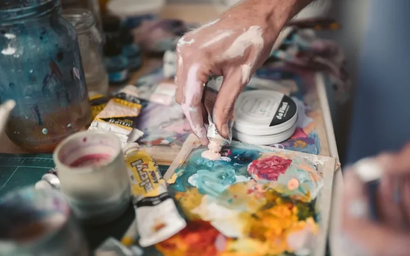 A detailed view of an artist's hand mixing vivid oil paints with a palette knife.
