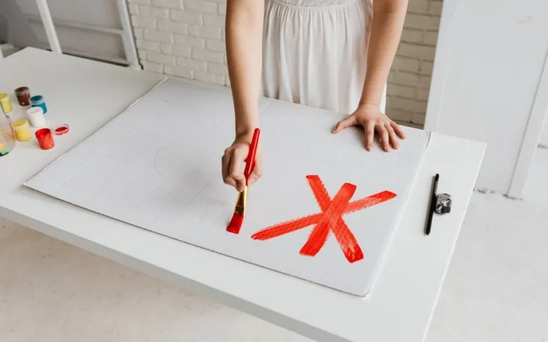 A woman in a white dress paints a red X on cardboard inside a sunlit studio.