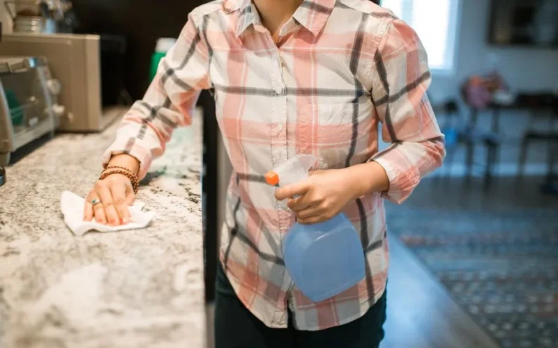 A woman wearing a plaid shirt cleans a kitchen counter using a spray bottle and cloth.