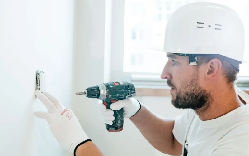 Side view of a construction worker using a drill on a wall, wearing safety gear.