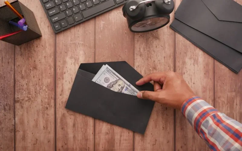 A hand placing a 100 dollar bill in a black envelope on a wooden desk with stationery, keyboard, and alarm clock.