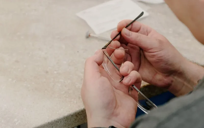From above of anonymous male master sitting at table and fastening temple with screw while repairing eyeglasses in professional workshop