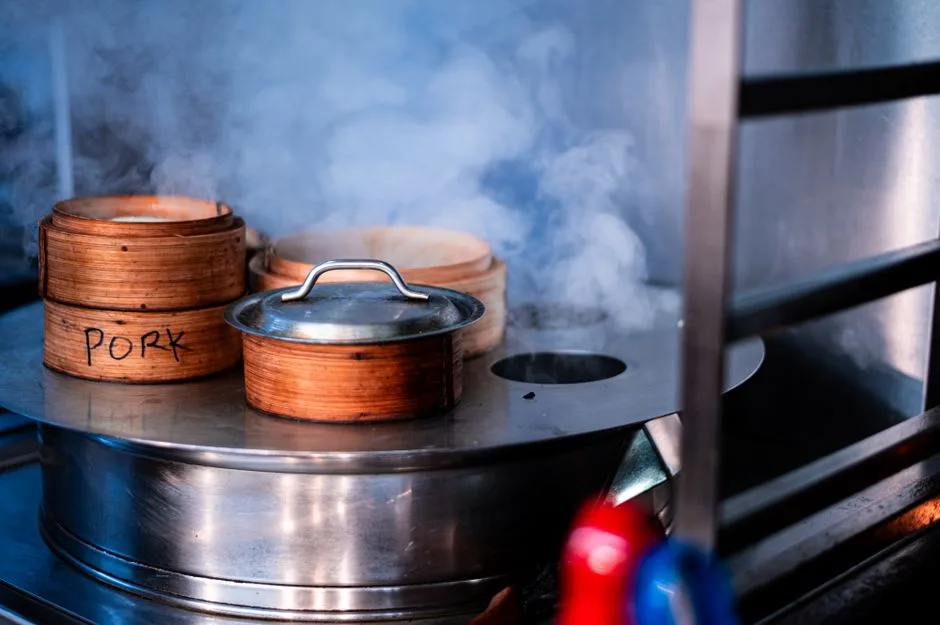 Wooden steamer baskets labeled 'Pork' steaming on a stovetop, creating aromatic vapor.