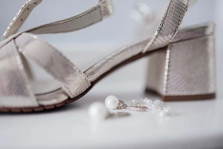 Close-up of bridal block heel sandals paired with pearl earrings on a white surface.