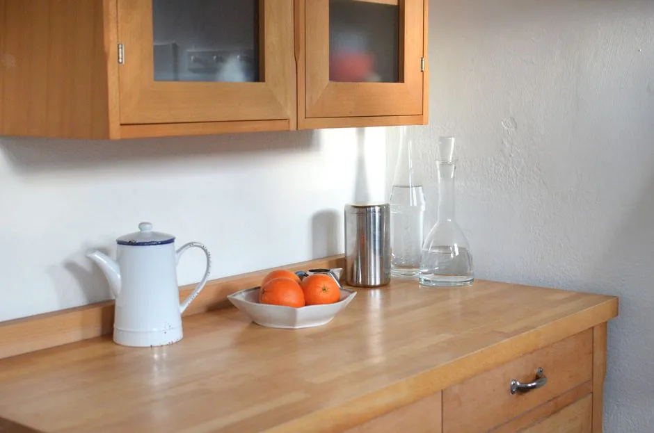 Wooden cabinet for dishes placed above wooden table with teapot glassware and bowl of oranges in modern light kitchen